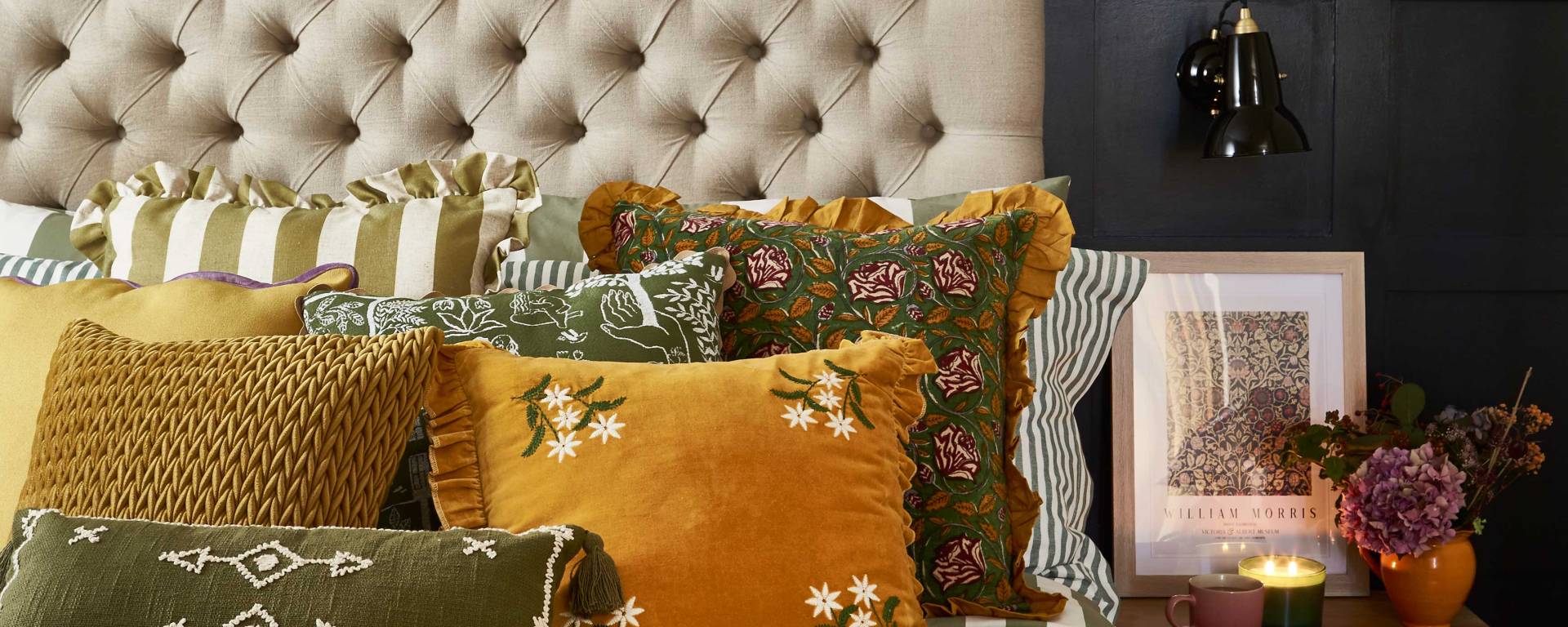 A cosy bedroom featuring a tufted beige headboard against dark panelling, styled with layered cushions in mustard yellow, olive green, and patterned fabrics. A striped duvet and floral throw add texture, while a bedside table with a black lamp, framed artwork, and lit candles create a warm, autumnal atmosphere.