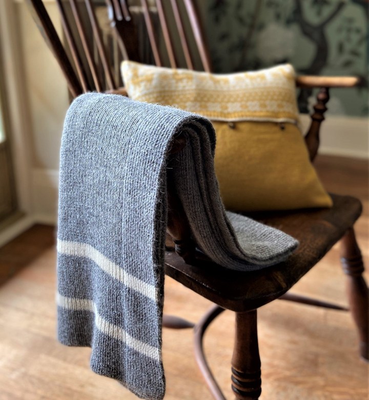 Grey ribbed knitted throw with white stripes draped over a dark wooden armchair with a mustard cushion.
