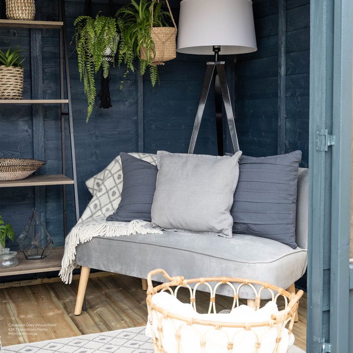 Cosy garden room interior with dark blue timber walls, a grey cushioned sofa, hanging plants, open shelving and a tall floor lamp.