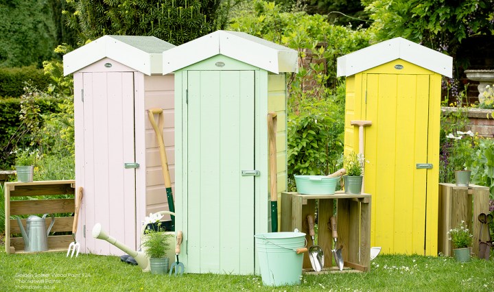 Three small wooden garden sheds painted pale pink, mint green and bright yellow, surrounded by gardening tools, pots and watering cans in a lush garden.