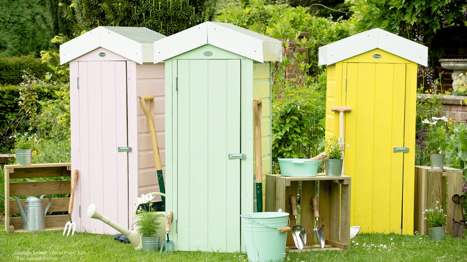 Three small wooden garden sheds painted pale pink, mint green and bright yellow, surrounded by gardening tools, pots and watering cans in a lush garden.
