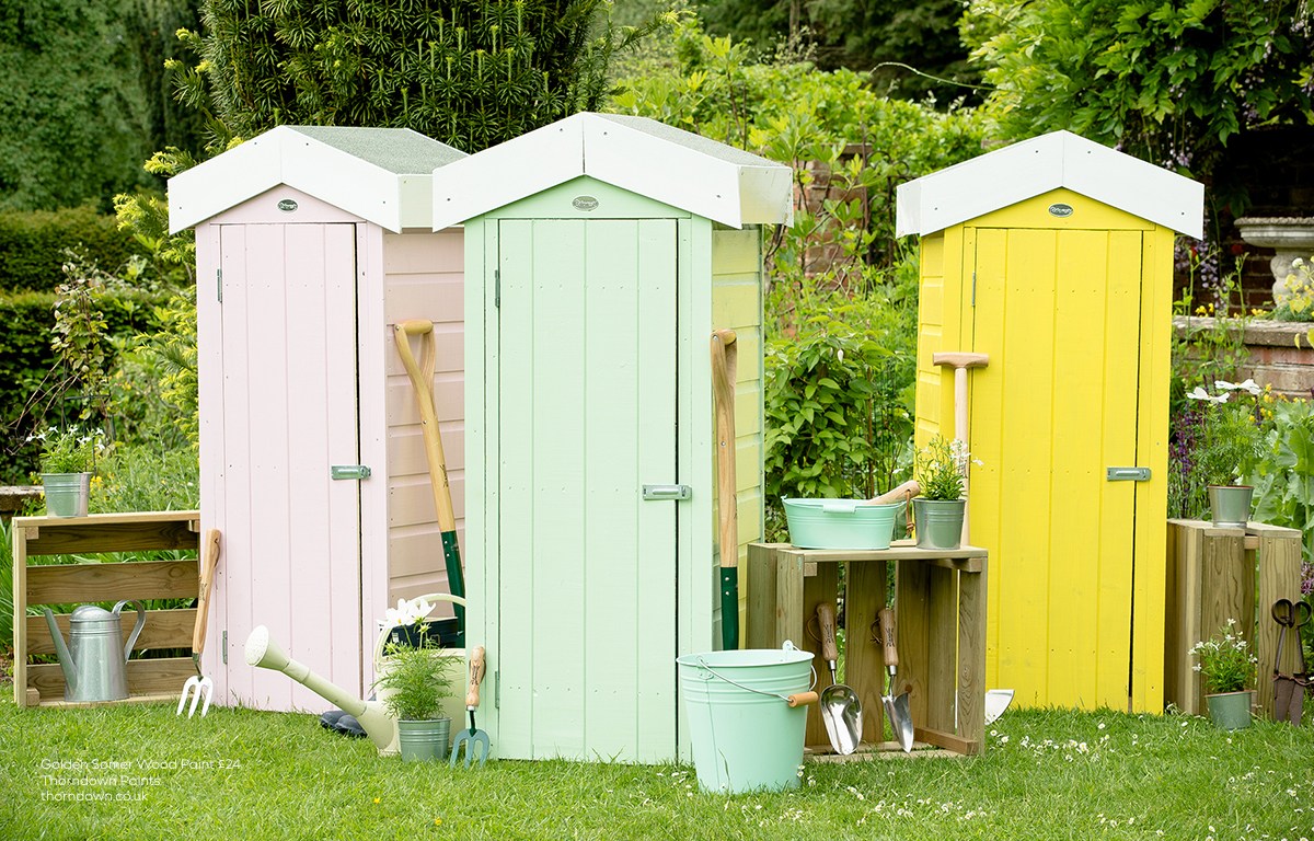 Three small wooden garden sheds painted pale pink, mint green and bright yellow, surrounded by gardening tools, pots and watering cans in a lush garden.