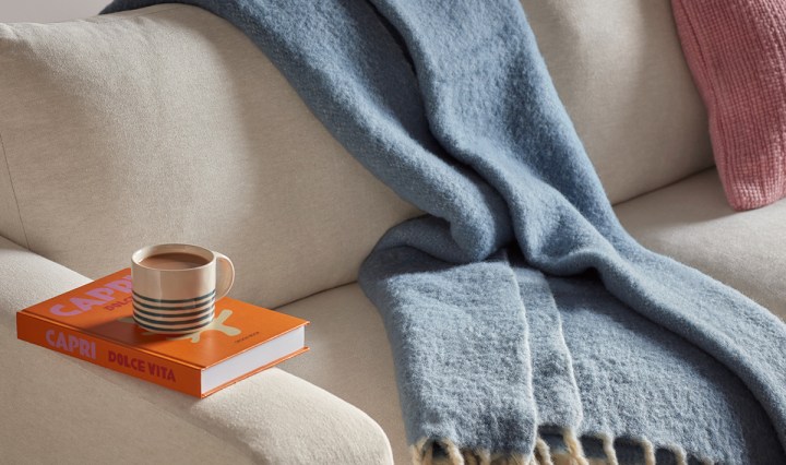 Blue fringed wool blanket draped over a cream sofa, styled with a striped mug on an orange coffee-table book and a pink cushion.