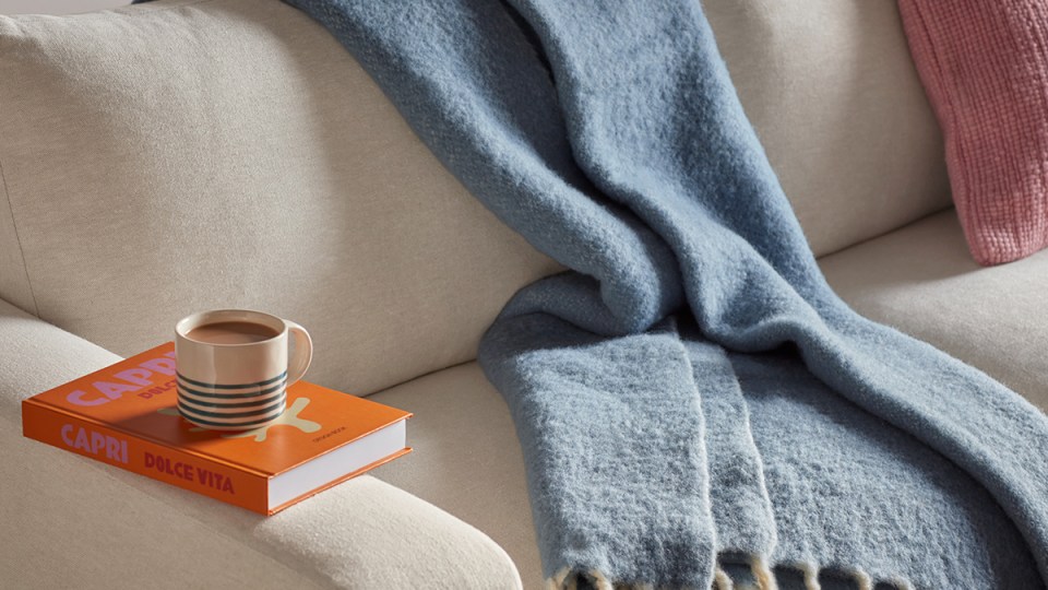 Blue fringed wool blanket draped over a cream sofa, styled with a striped mug on an orange coffee-table book and a pink cushion.