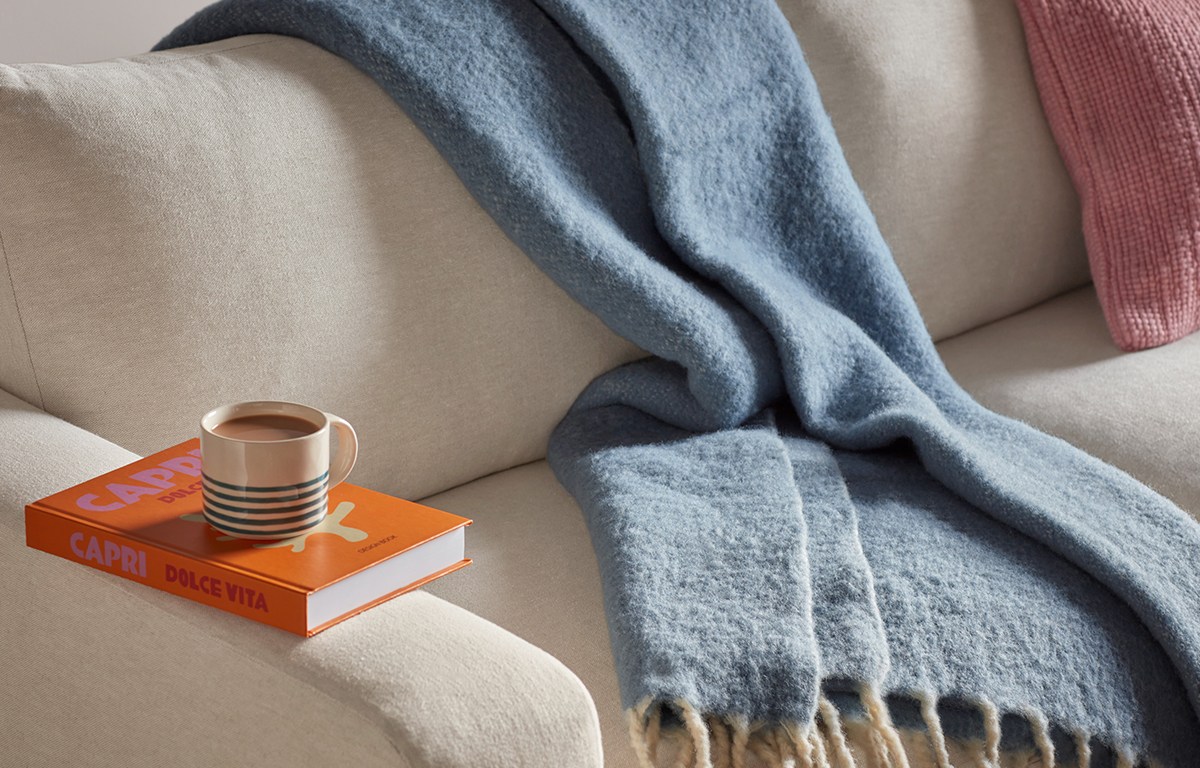 Blue fringed wool blanket draped over a cream sofa, styled with a striped mug on an orange coffee-table book and a pink cushion.