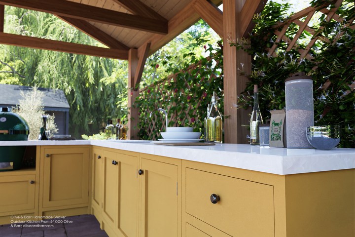 Covered outdoor kitchen with mustard-yellow Shaker cabinetry, white countertops, a green ceramic grill, sink, and climbing plants on a wooden trellis.