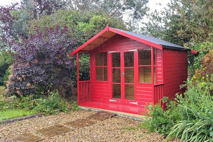 Bright red timber garden shed with glazed double doors and windows, set among dense, mature planting in a leafy garden.